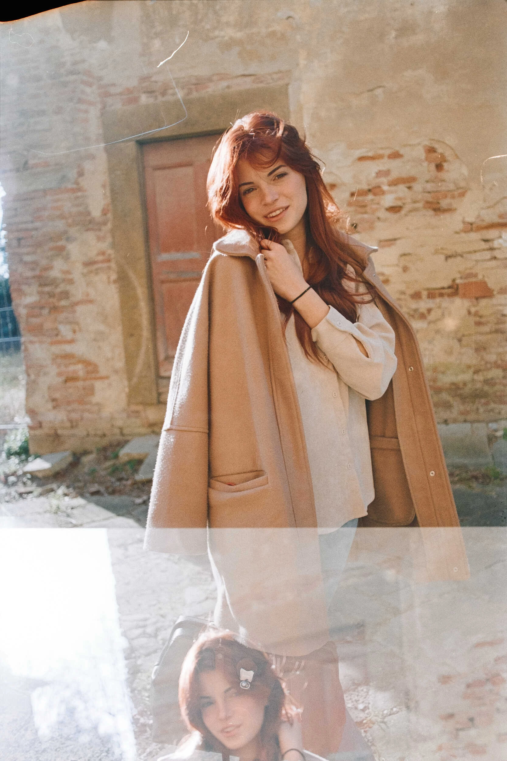 Red-haired model in a camel coat smiling by a brick wall, analog film portrait in Tuscany