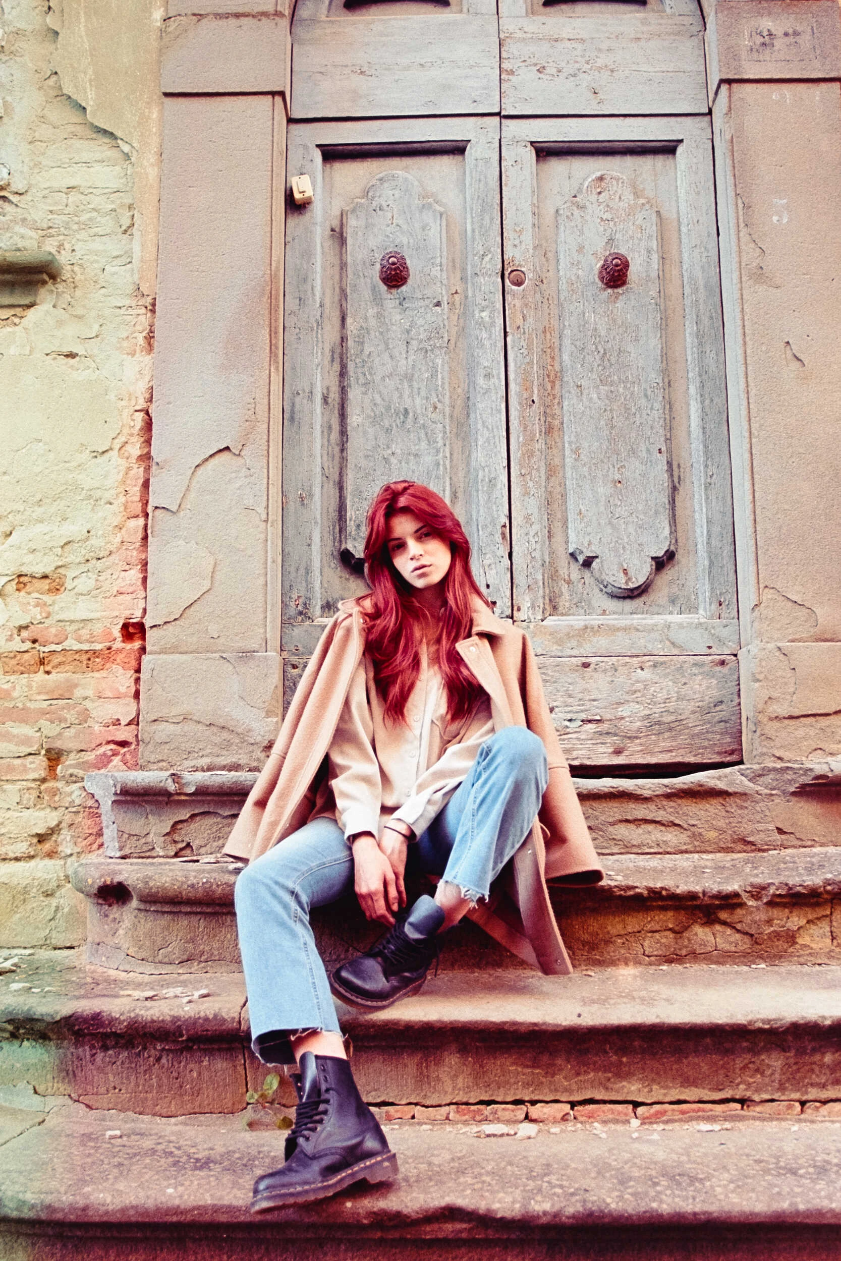 Red-haired model seated on steps in a camel coat and jeans, analog film portrait in Tuscany