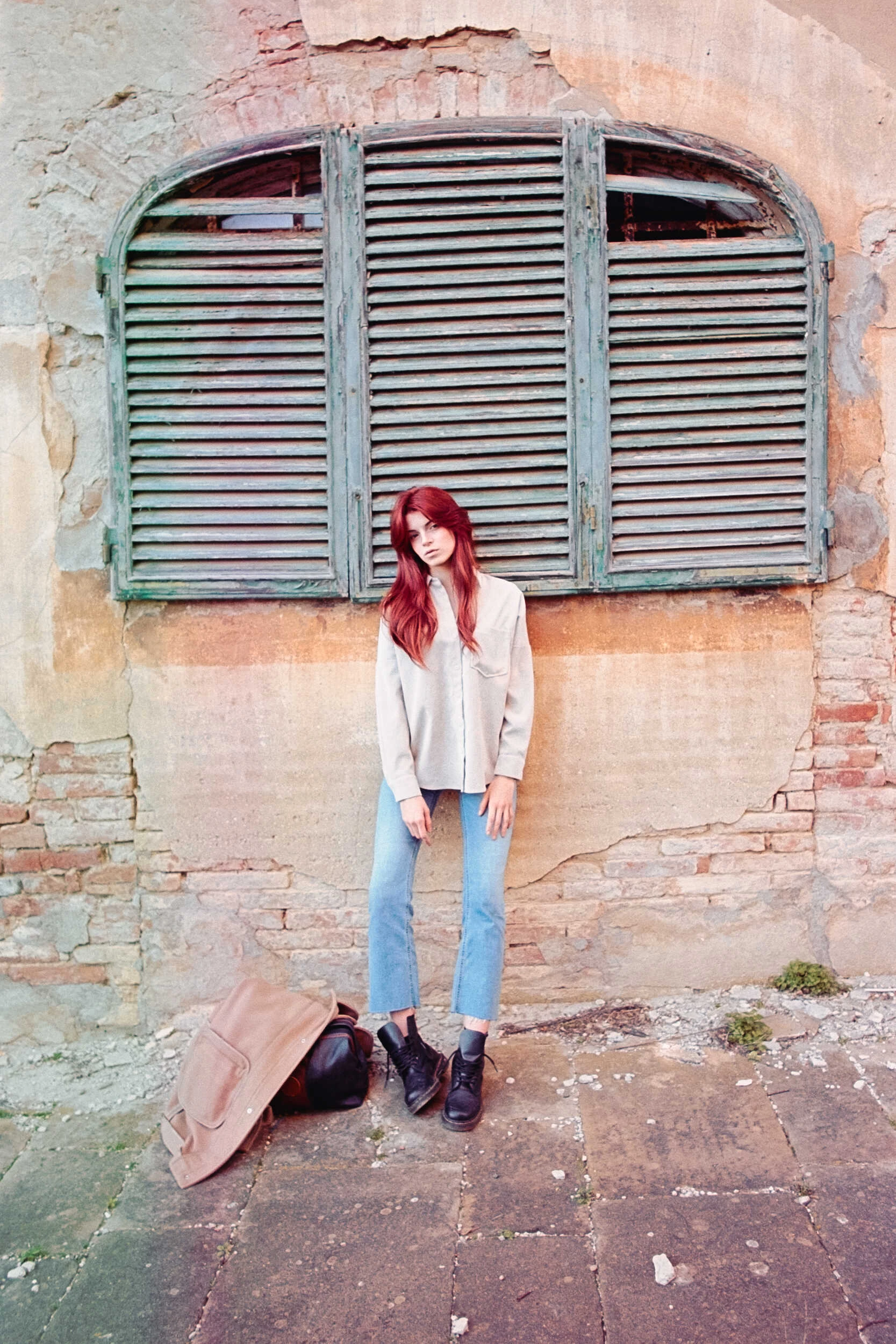 Red-haired model in a cream shirt and jeans by an old window, analog film portrait in Tuscany