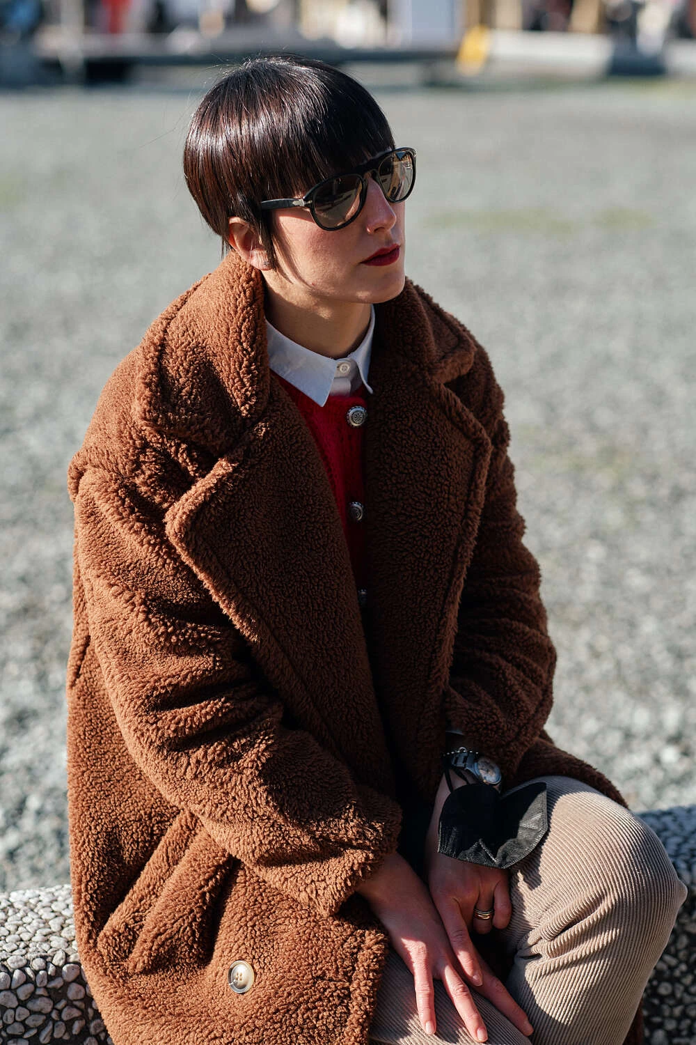 Person in a brown teddy coat and sunglasses seated, street style at Pitti Uomo 101 in Florence