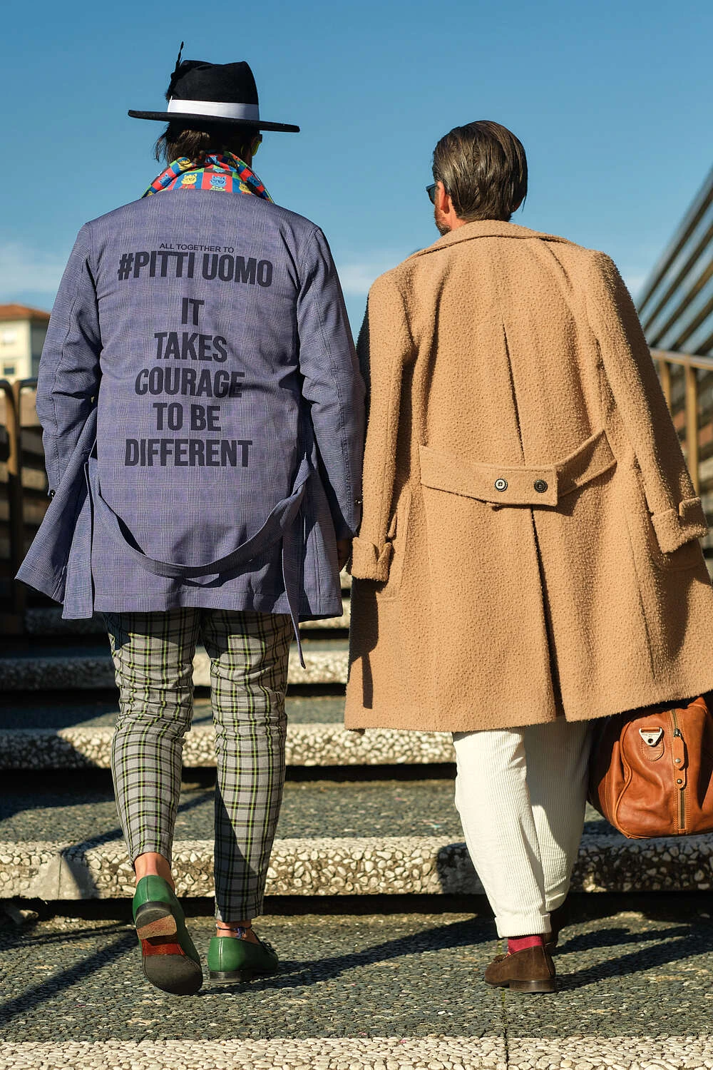 Man in a Pitti Uomo slogan jacket seen from behind beside a camel coat, street style at Pitti Uomo 101 in Florence