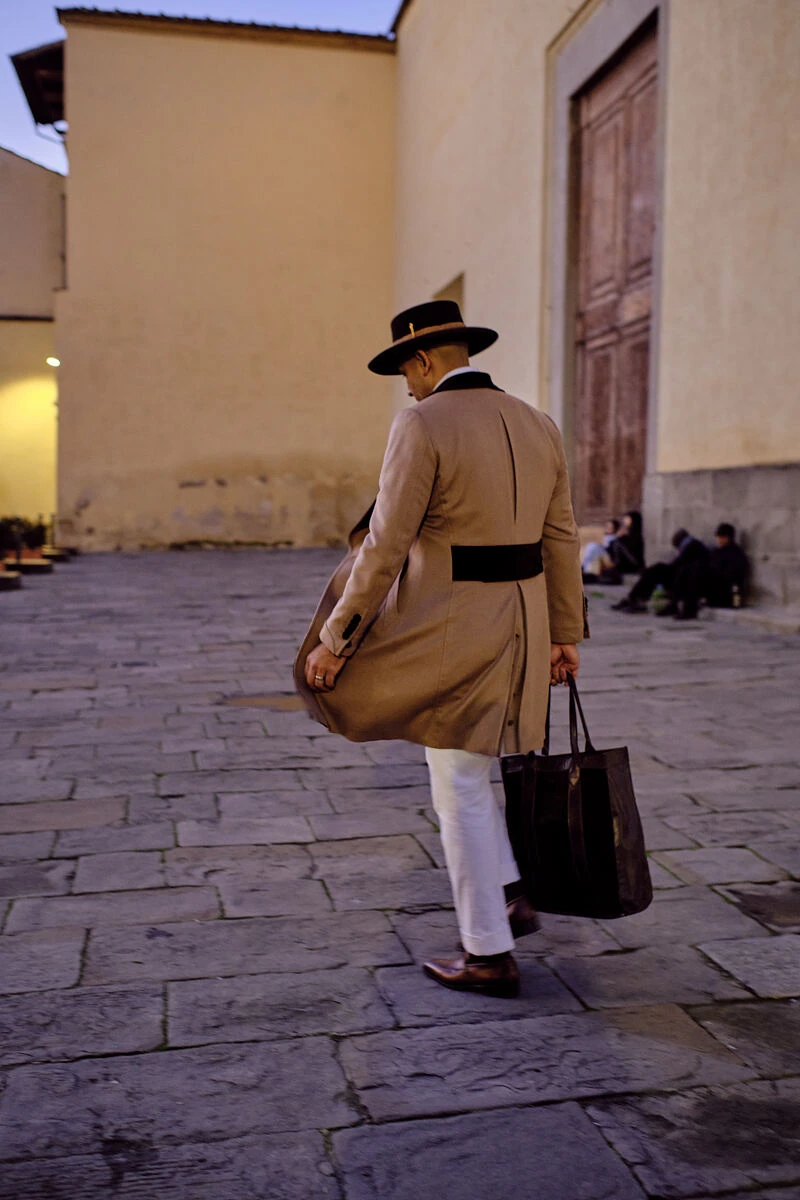 Man in a tan overcoat and hat walking a Florence street with a tote bag, male fashion street style