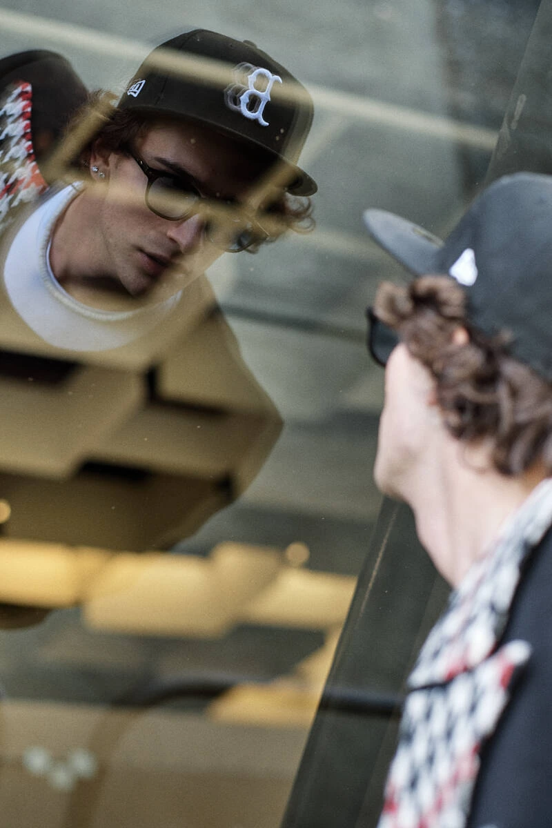 Man with a cap and sunglasses reflected in glass, street style at Pitti Uomo 103 in Florence