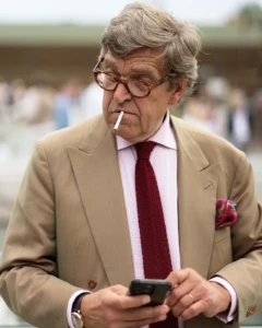 Older man in a tan suit with a red knit tie smoking a cigarette, street style at Pitti Uomo 104 in Florence