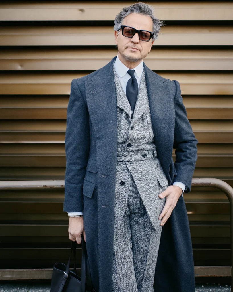 Man in a grey herringbone double-breasted suit and navy overcoat with sunglasses, street style at Pitti Uomo 105 in Florence