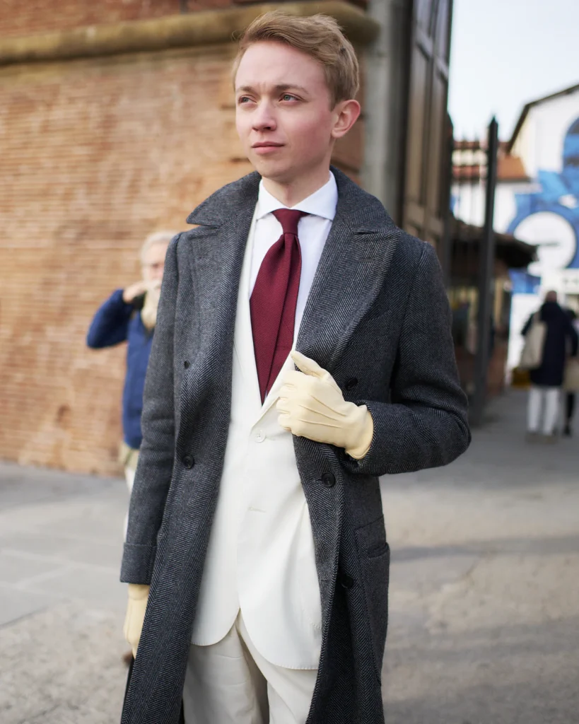 Blond man in a grey overcoat and white suit holding cream gloves, street style at Pitti Uomo 105 in Florence