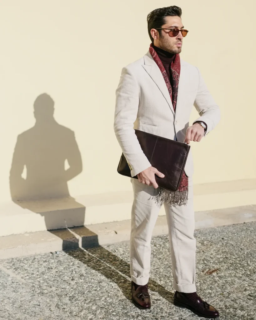 Man in a cream suit with a burgundy scarf holding a leather clutch, street style at Pitti Uomo 105 in Florence