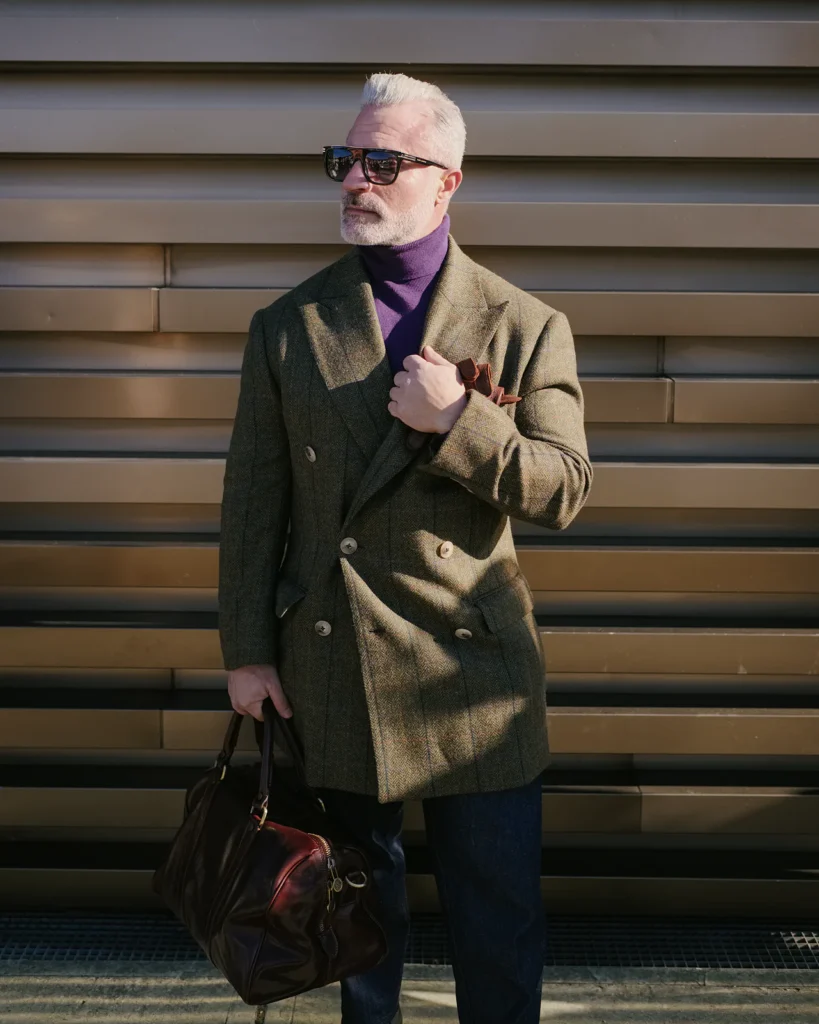 Grey-haired man in an olive double-breasted tweed coat over a purple turtleneck, street style at Pitti Uomo 105 in Florence