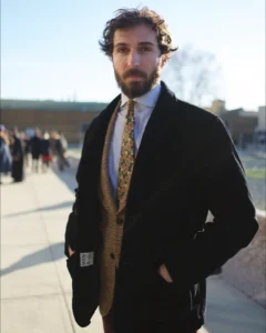 Bearded man in a dark overcoat over patterned knitwear, street style at Pitti Uomo 105 in Florence