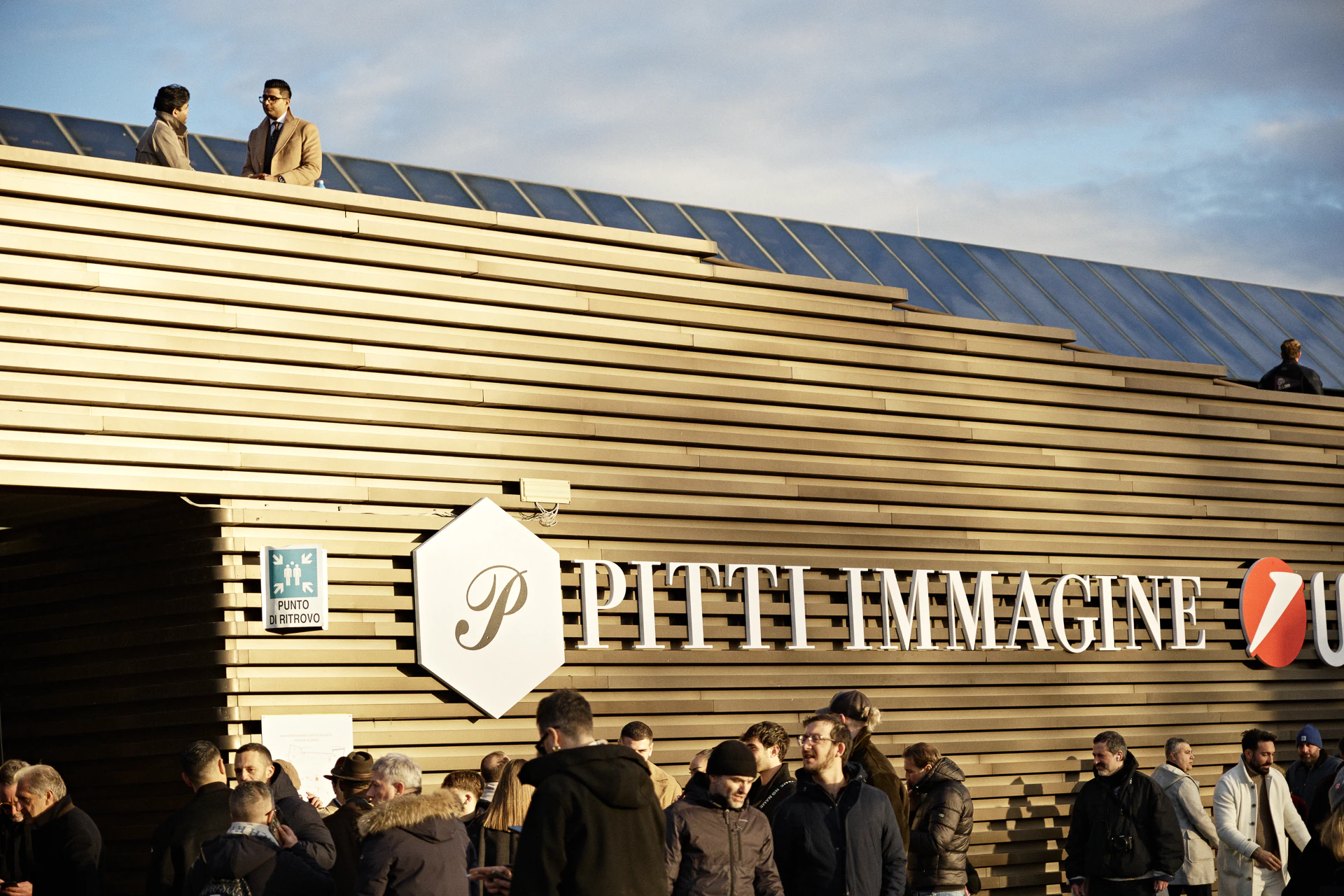Golden slatted-wood entrance pavilion of Pitti Immagine Uomo 107 at Fortezza da Basso, Florence, with attendees gathering at dusk — menswear trade fair coverage.