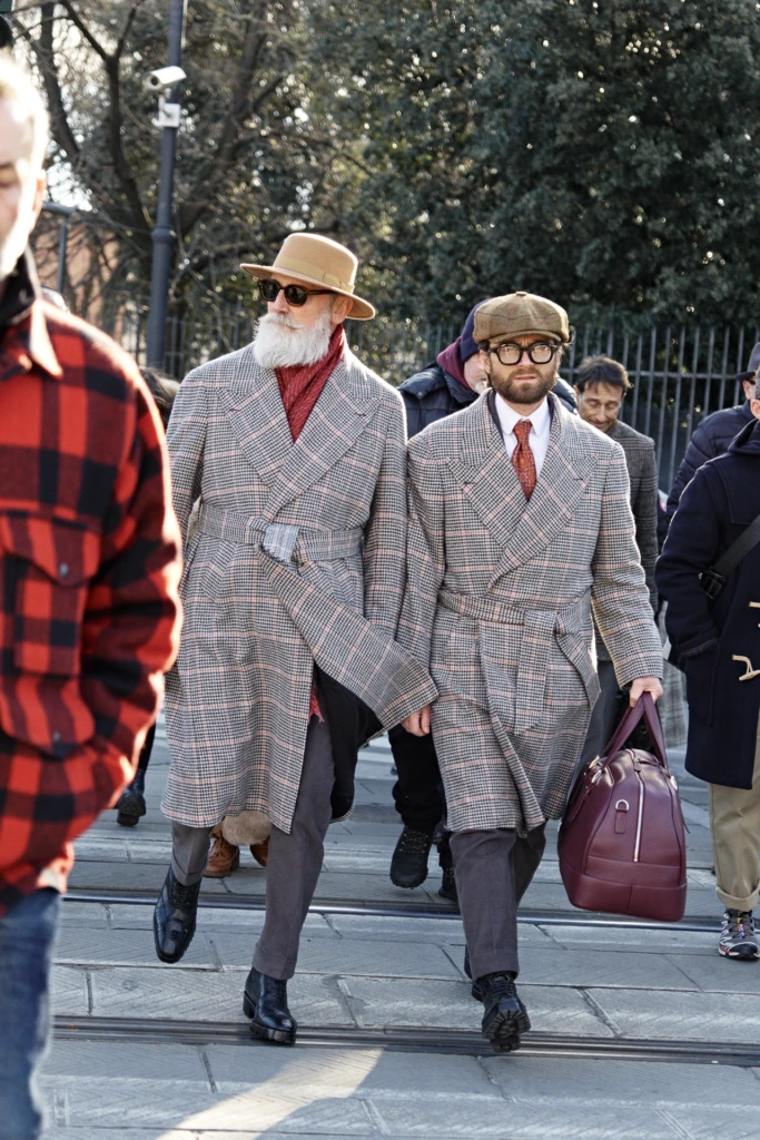 Two men in matching grey houndstooth belted overcoats — one with a tan fedora and red scarf — walking to Pitti Immagine Uomo 107, Florence menswear street style.