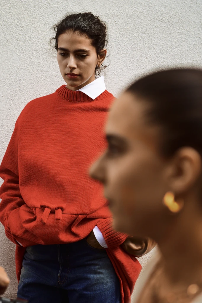 Woman in an oversized rust-orange sweater with white shirt collar peeking out, hands in pockets, another guest with a gold hoop earring blurred in the foreground, Milano Fashion Week street style