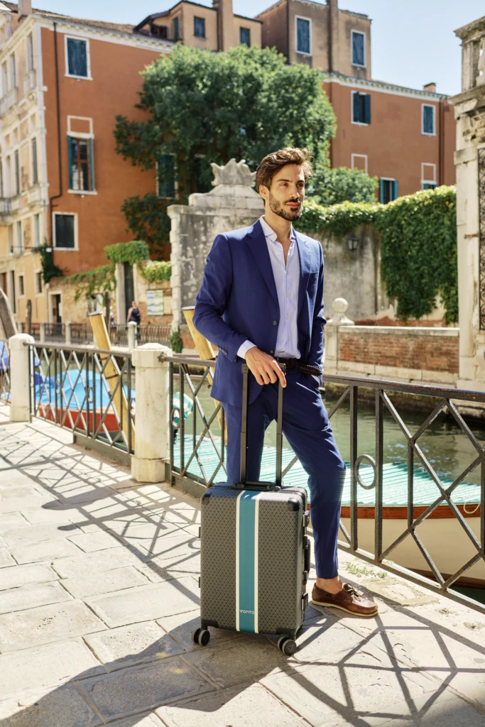 Man in navy suit with a Vonto striped hard-shell rolling suitcase beside a Venice canal bridge
