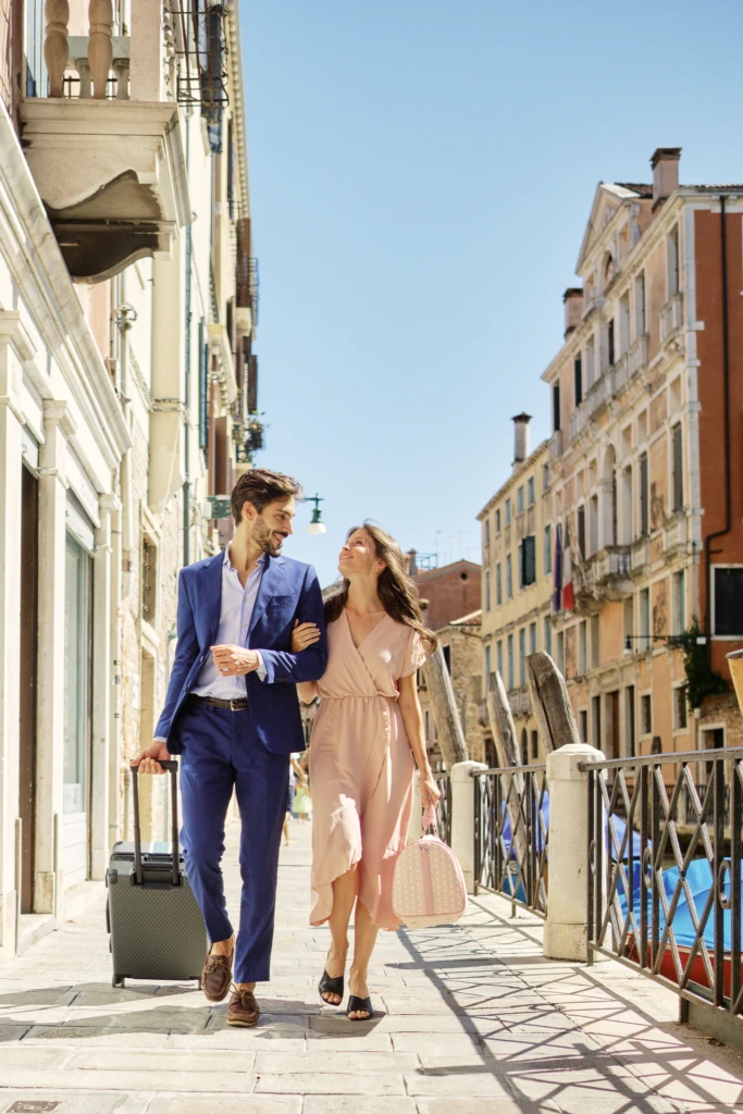 Couple walking a sunlit Venice alley with a Vonto rolling travel suitcase and pink shopping tote