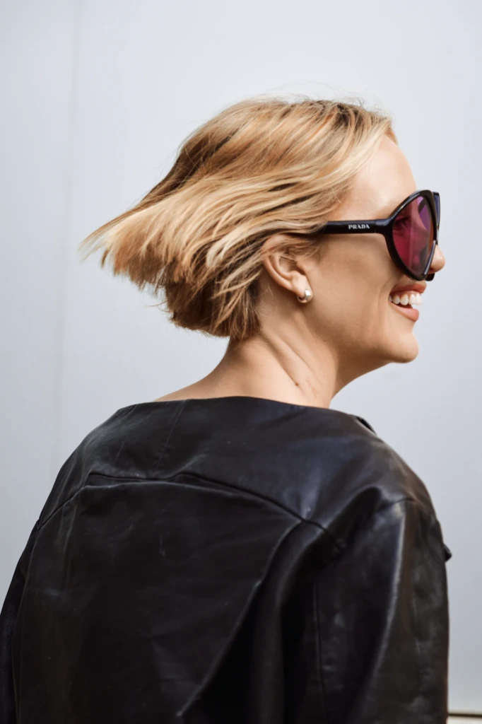 Woman with a short blonde bob mid-flip in the wind, wearing round pink-lensed Prada sunglasses and a black leather jacket, laughing, Milano Fashion Week street style