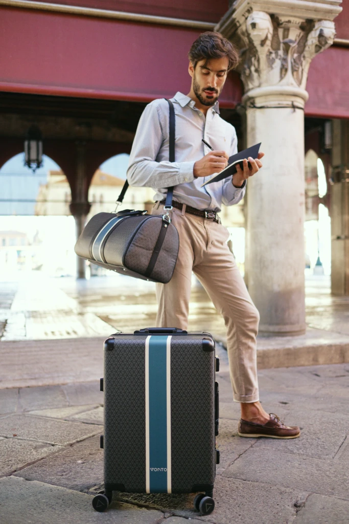 Male model with a Vonto leather duffel and monogram rolling suitcase in a sunlit Venetian piazza
