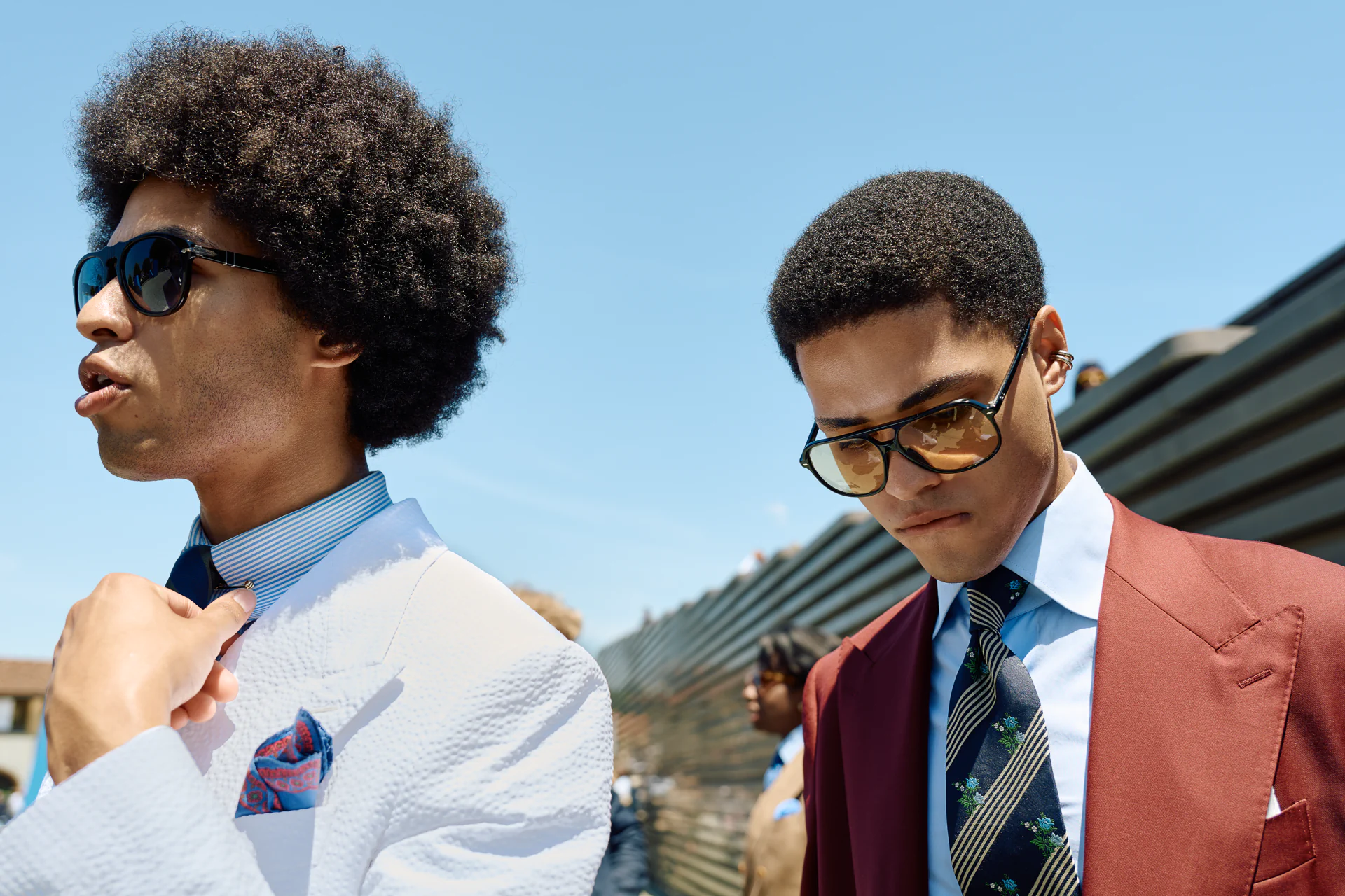 Two men in sunglasses at Pitti Immagine Uomo 110, one in a white seersucker jacket with a paisley pocket square, the other in a rust suit with a striped floral tie