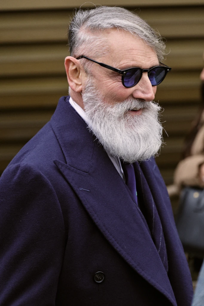 Elderly man in profile wearing round black sunglasses and a navy overcoat with a boutonniere, Pitti Immagine Uomo 109 fair