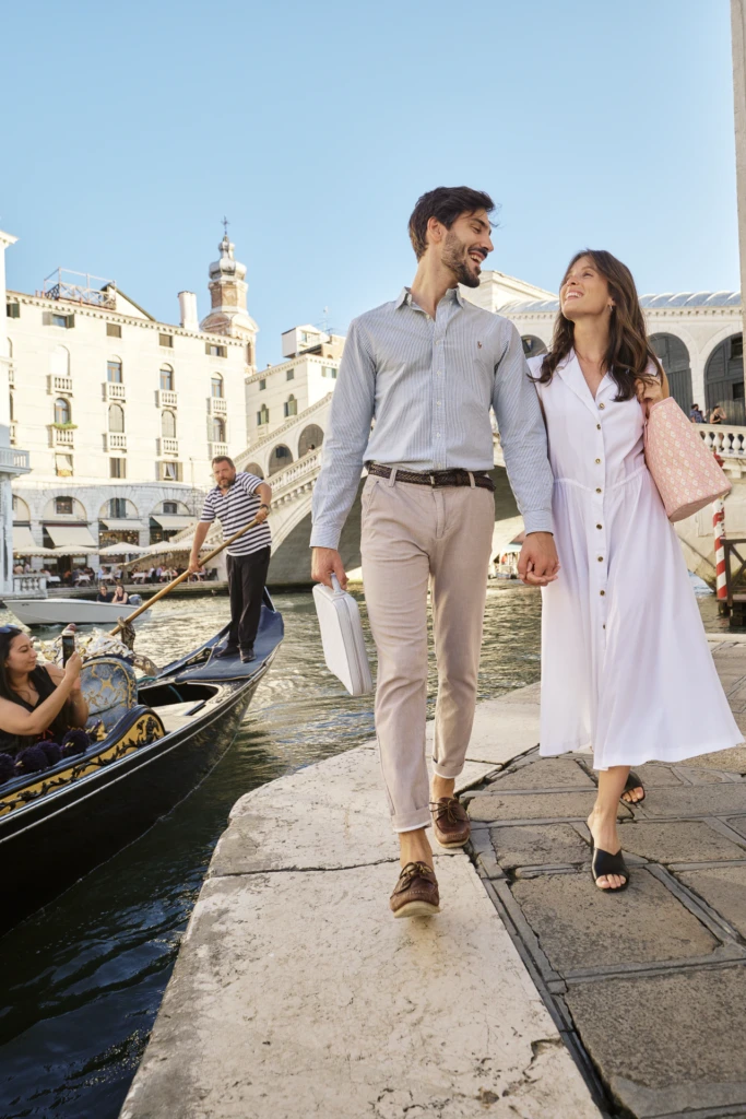 Elegant couple walking a Venice canal quay past a gondola with a Vonto leather bag in hand