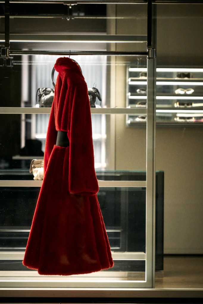 Long red faux-fur coat displayed on a hanger inside a boutique shop window, sunglasses visible on shelves behind