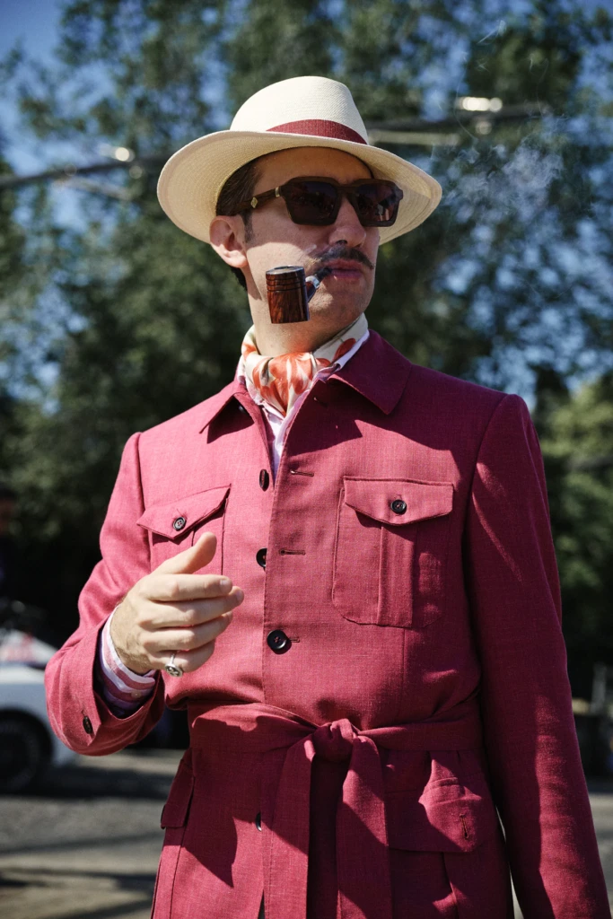 Man in a burgundy belted suit smoking a pipe, wearing a cream straw fedora with a red band at Pitti Uomo
