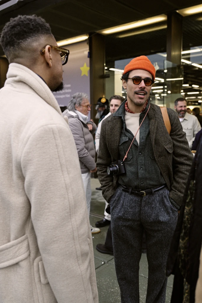 Two men talking, one in an orange beanie with a camera around his neck and an olive tweed jacket, Pitti Immagine Uomo 109 fair, Florence