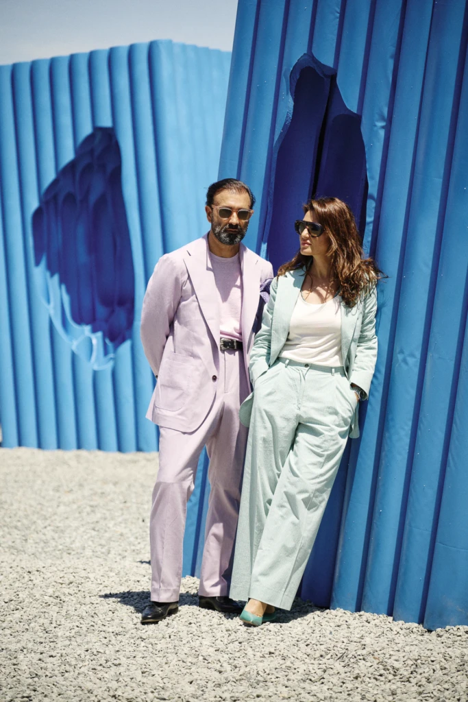 Couple against a blue corrugated wall, man in a lilac suit and woman in a mint suit, both in sunglasses