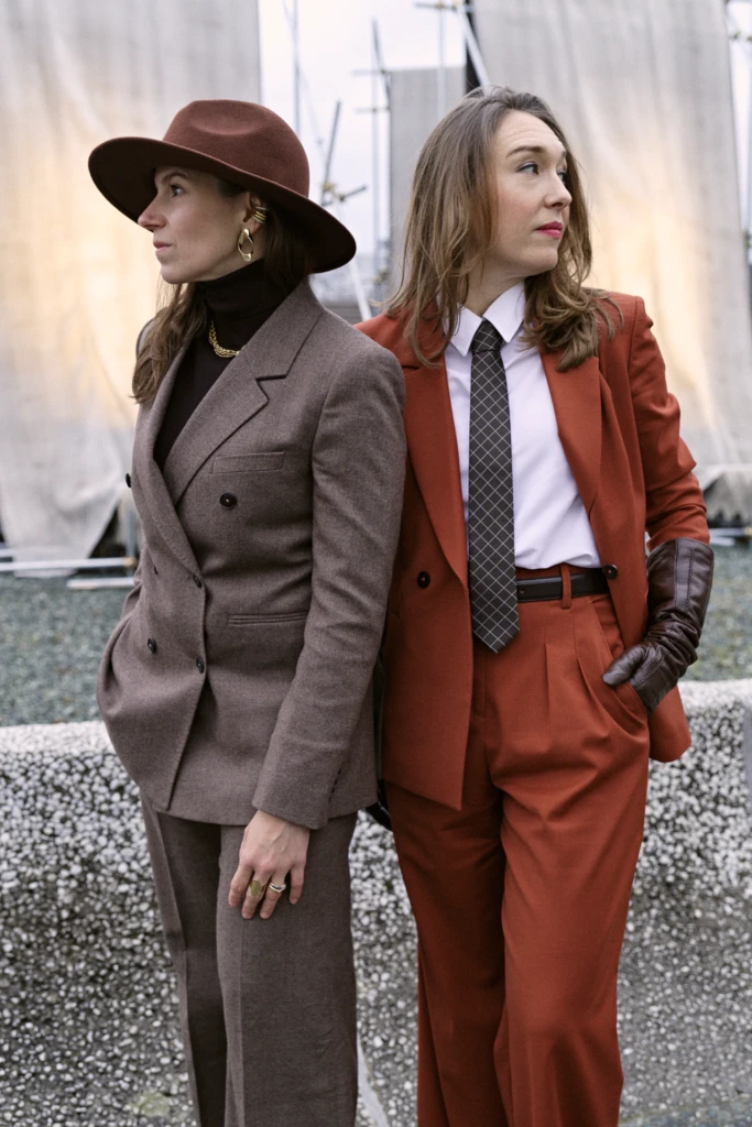 Two women in tailored suits, one in brown with a wide-brim hat, the other in a rust suit with a check tie, Pitti Immagine Uomo 109 fair