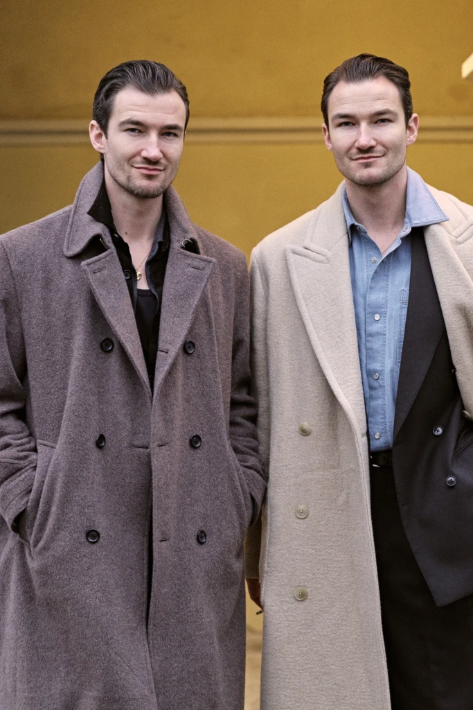 Two young men in wool overcoats, one taupe and one cream, smiling together at the Pitti Immagine Uomo 109 fair, Florence