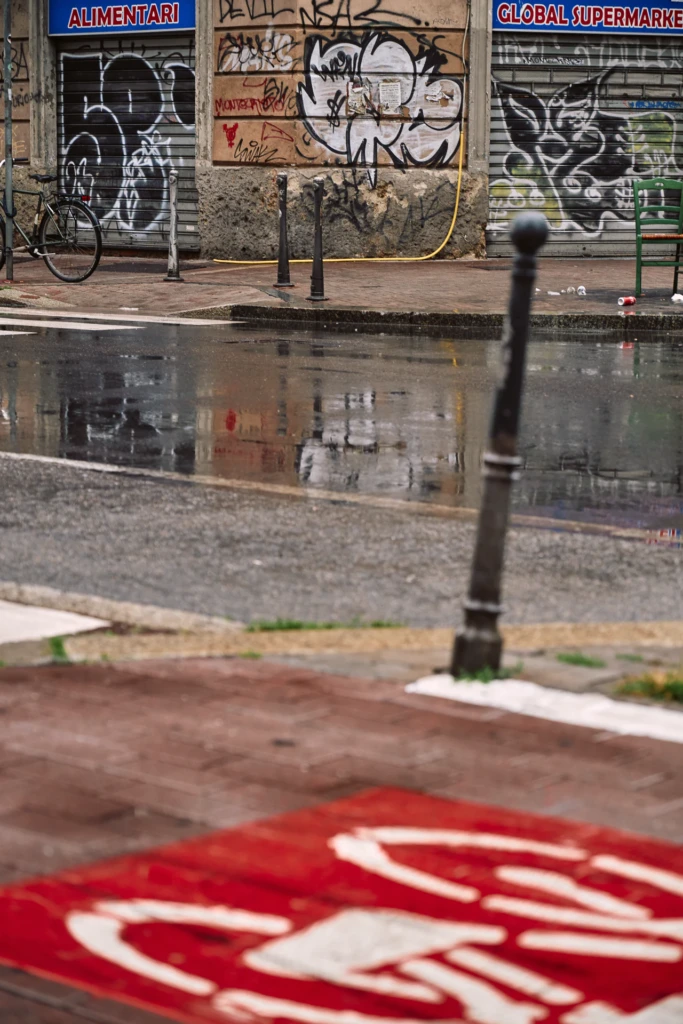 Rainy Milano street corner with graffiti-covered shop shutters, a parked bicycle, and reflections on wet pavement