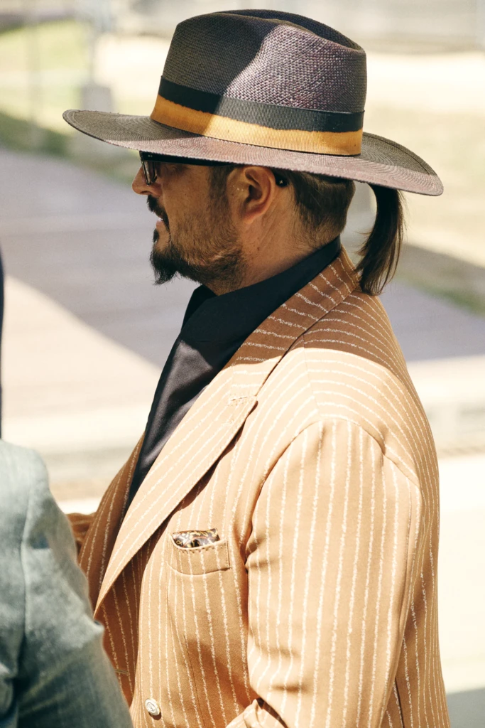 Man wearing a wide-brim striped hat with a black and gold band and a tan pinstripe suit, hair in a ponytail
