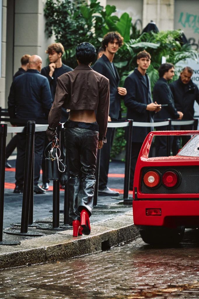 Rear view of a model in a cropped brown leather jacket and black leather trousers with red patent heels walking past a red Ferrari F40 on a rainy Milano street, onlookers gathered behind barriers