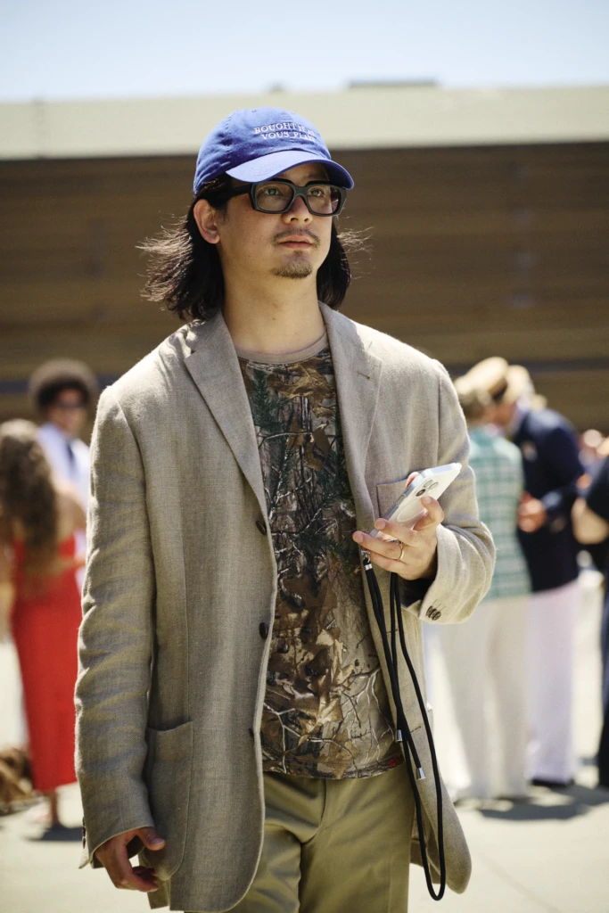 Man in a blue baseball cap and camouflage t-shirt under a beige blazer at Pitti Uomo