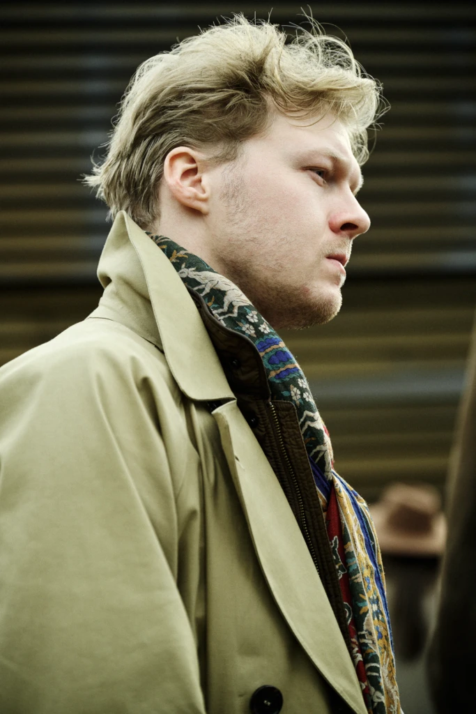 Blond man in profile wearing a khaki trench coat over a green and gold paisley scarf, Pitti Immagine Uomo 109 fair, Florence