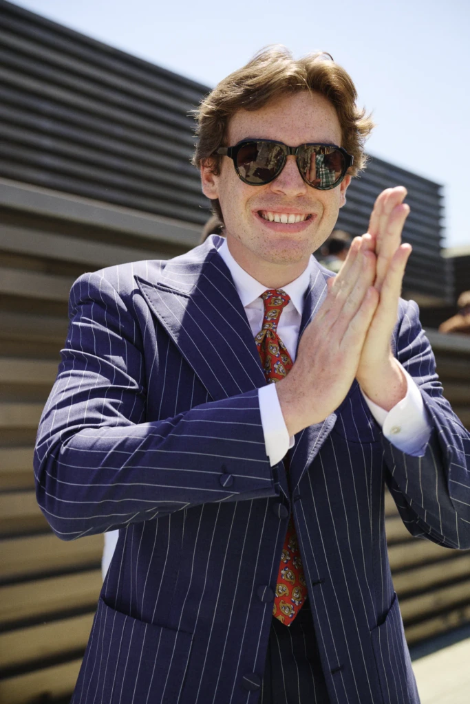 Man in a navy pinstripe suit and orange paisley tie clapping his hands, wearing sunglasses at Pitti Immagine Uomo 110