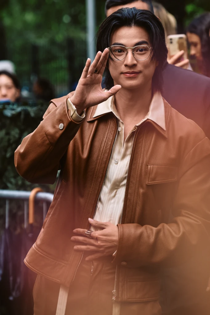 Man with shoulder-length dark hair wearing round wire-frame glasses and a tan leather jacket over a cream shirt, adjusting his glasses, Milano Fashion Week street style