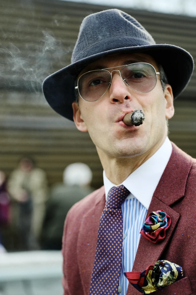 Man in a grey fedora smoking a cigar, wearing aviator sunglasses and a burgundy tweed suit with a floral boutonniere, Pitti Immagine Uomo 109 fair
