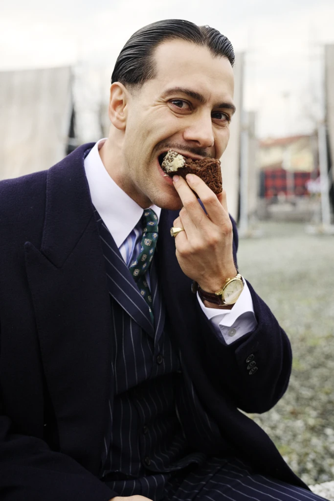 Man in a navy pinstriped suit and green tie eating a piece of cake, wearing a gold signet ring, Pitti Immagine Uomo 109 fair, Florence