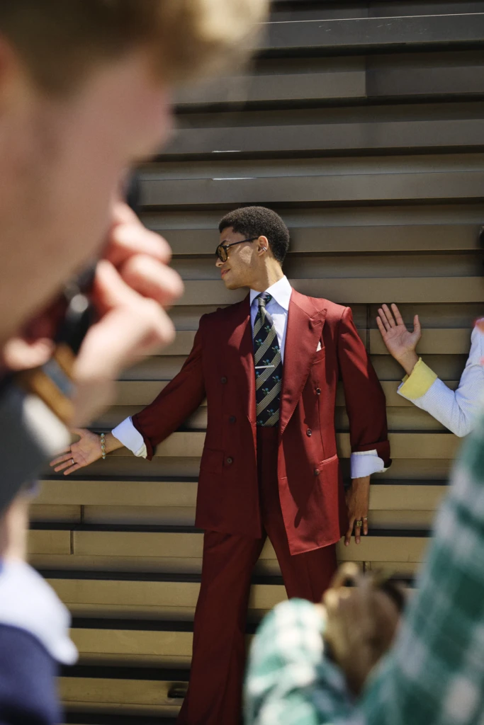 Man in a burgundy double-breasted suit with a striped patterned tie and tie bar, arms outstretched, wearing sunglasses