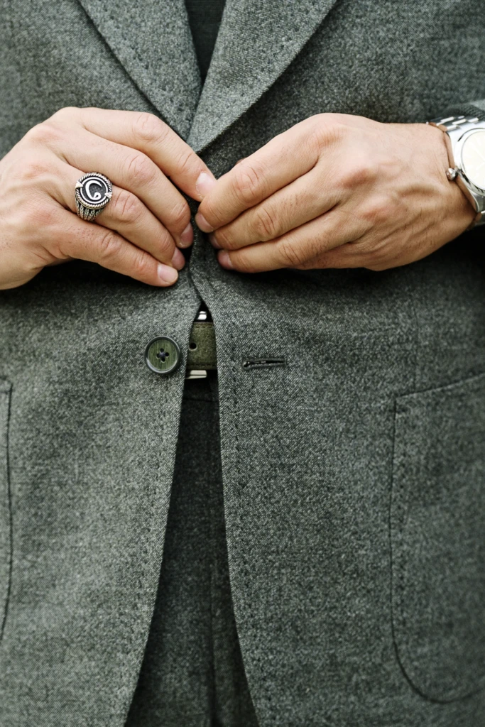 Close-up of hands buttoning a grey tweed blazer, with a signet ring and steel watch, Pitti Immagine Uomo 109 fair, Florence