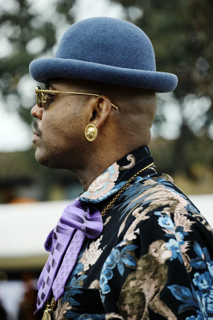 Man in profile wearing a blue bowler hat, gold earring and a floral print jacket with a purple bow tie, Pitti Immagine Uomo 109 fair, Florence