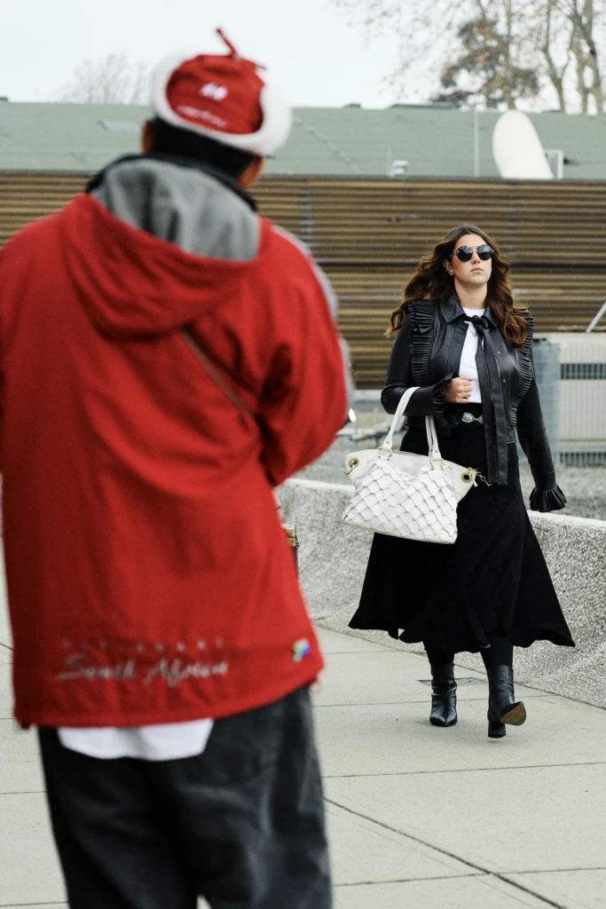 Woman in a black leather jacket with ruffled trim carrying a white woven handbag, walking past a man in a red hooded jacket, Pitti Immagine Uomo 109 fair