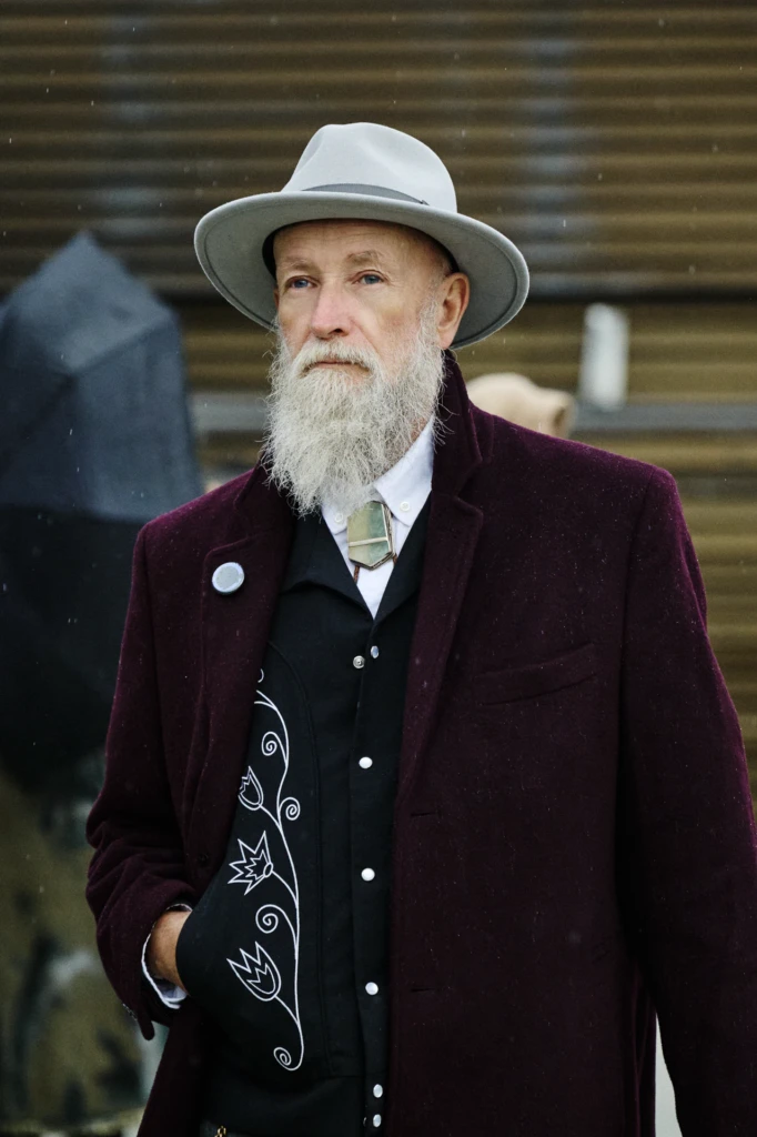 Elderly man in a grey wide-brim hat and maroon velvet coat with a bolo tie, wearing a black shirt with floral embroidery, Pitti Immagine Uomo 109 fair