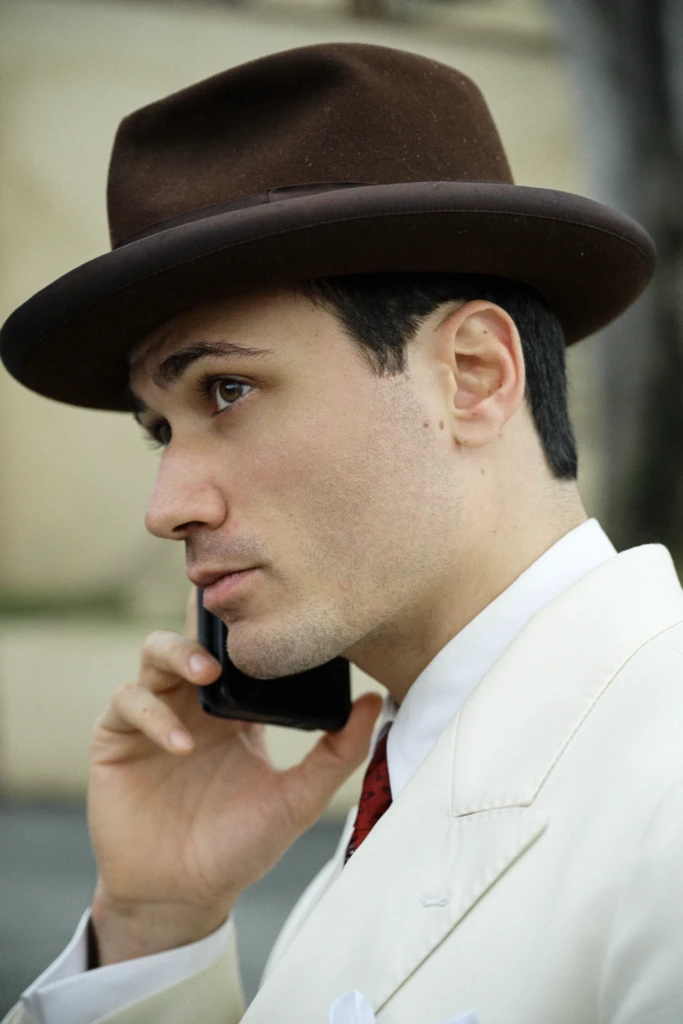 Young man in profile on the phone, wearing a brown fedora and cream double-breasted coat with a red tie, Pitti Immagine Uomo 109 fair, Florence