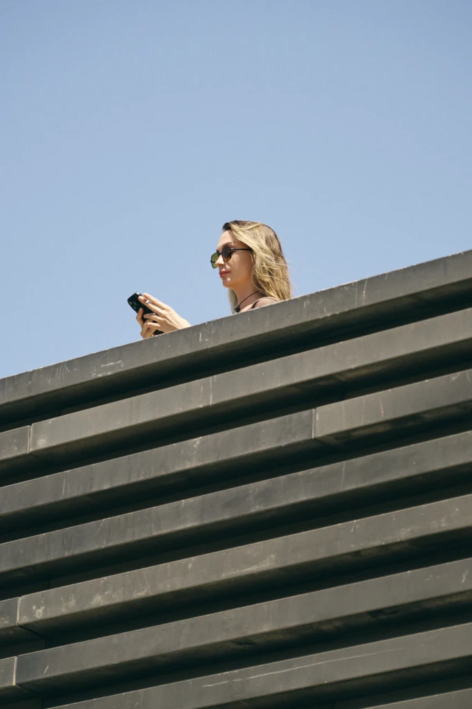 Woman leaning over a ledge checking her phone, wearing sunglasses