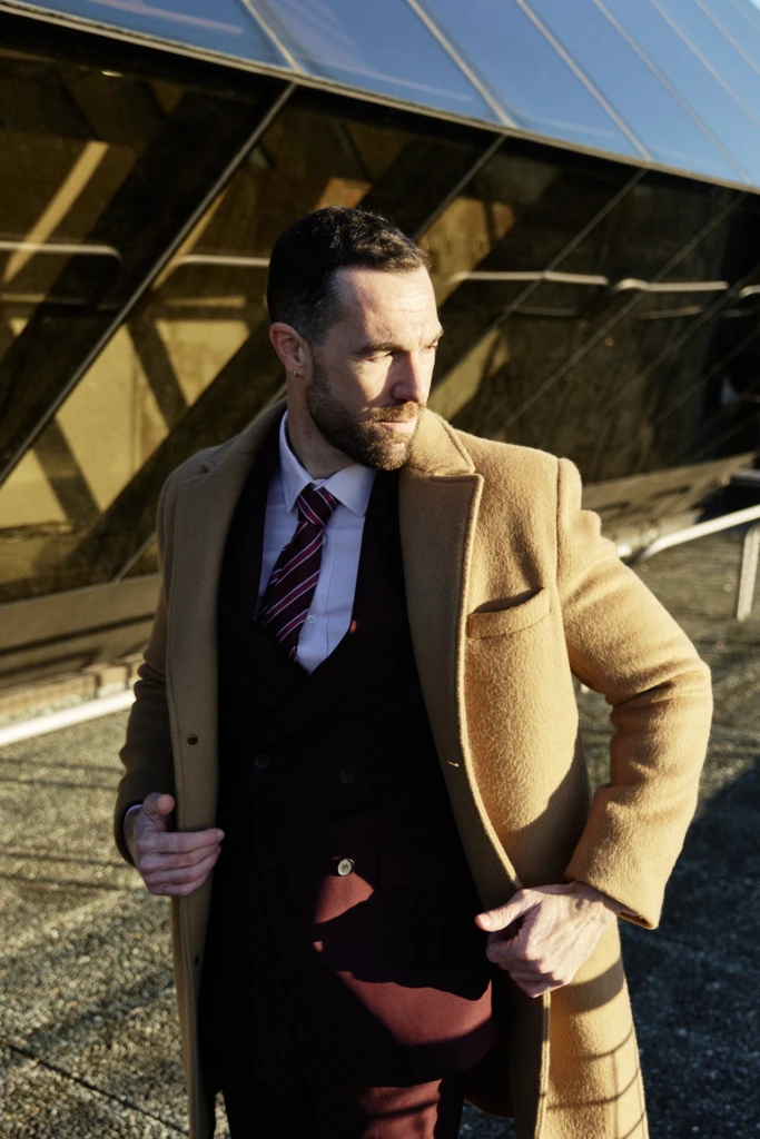 Hockerty editorial at Pitti Immagine Uomo 107 — camel double-breasted overcoat over burgundy suit against the fair's reflective glass architecture in Florence.