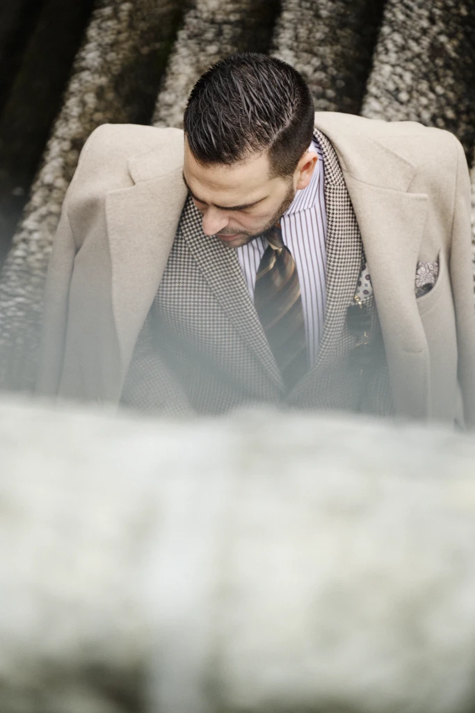 Man looking down as he buttons his houndstooth waistcoat and striped tie under a beige overcoat, Pitti Immagine Uomo 109 fair