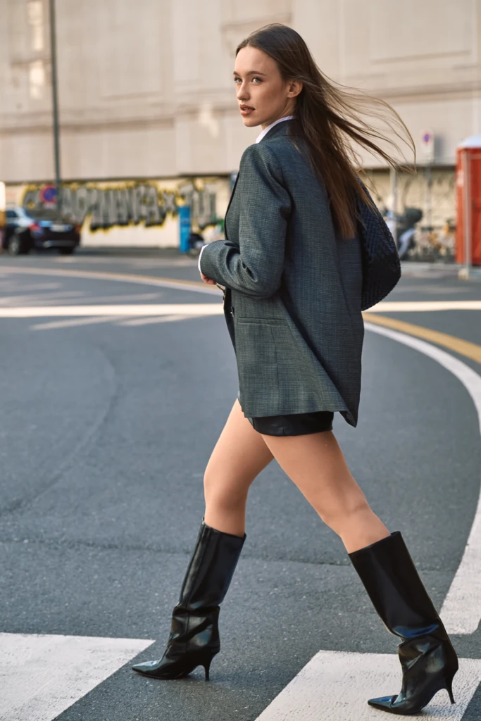 Woman in an oversized grey blazer, black leather shorts and knee-high black boots crossing a Milano street, looking back over her shoulder