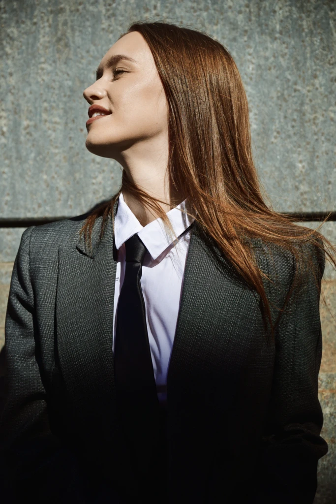 Profile portrait of a woman in a grey pinstripe blazer with white shirt and black tie, hair blowing in the wind, Milano Fashion Week street style