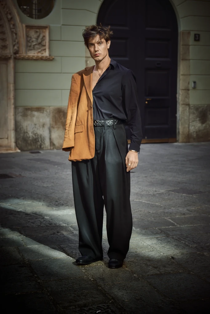 Marco Castelli in an oversized black shirt and wide-leg black trousers with a camel wool blazer draped over one shoulder, standing before a dark arched doorway, Milano Fashion Week street style